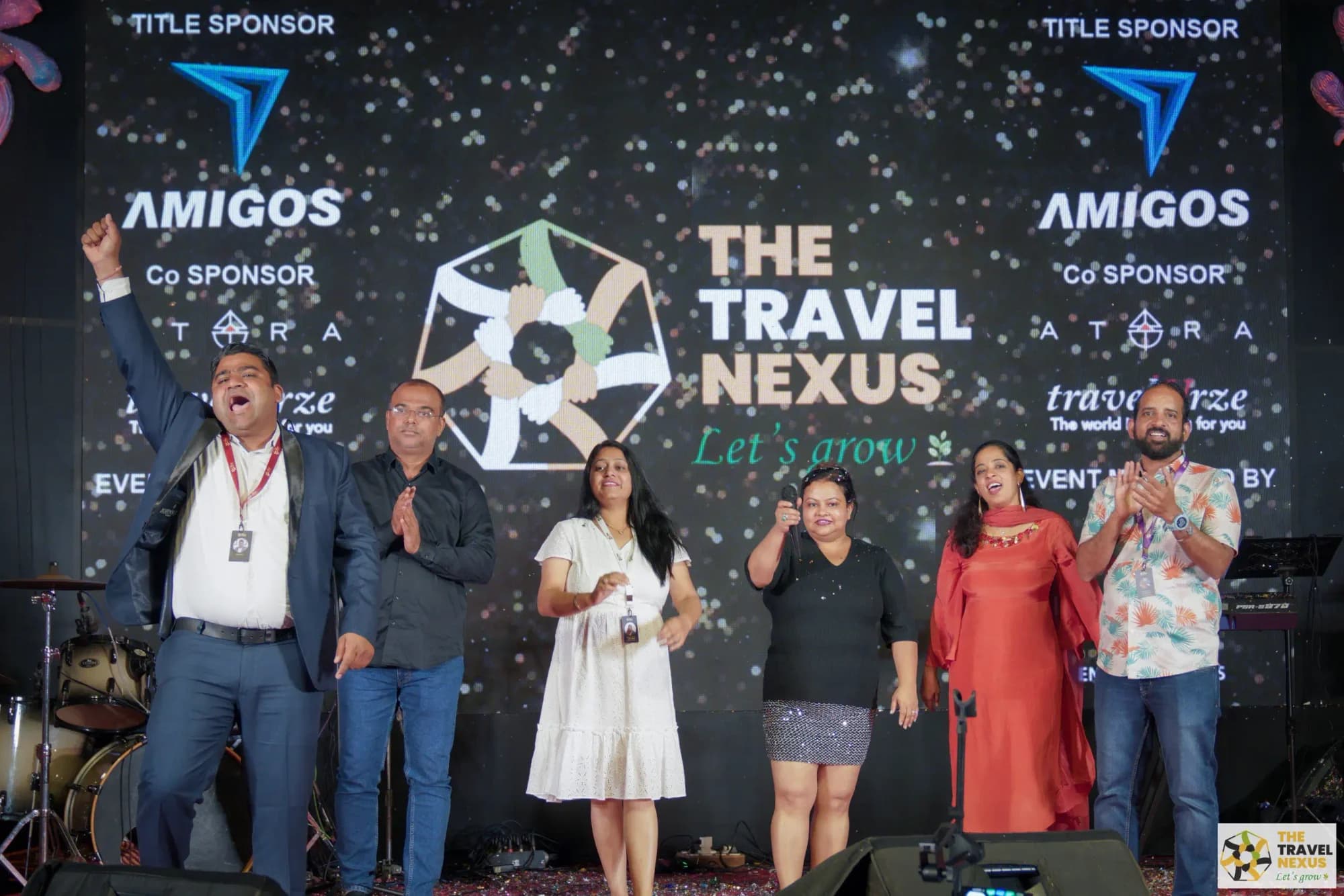 Six Travel Nexus members on stage, arms raised, in front of a large lit backdrop reading The Travel Nexus, Let's grow